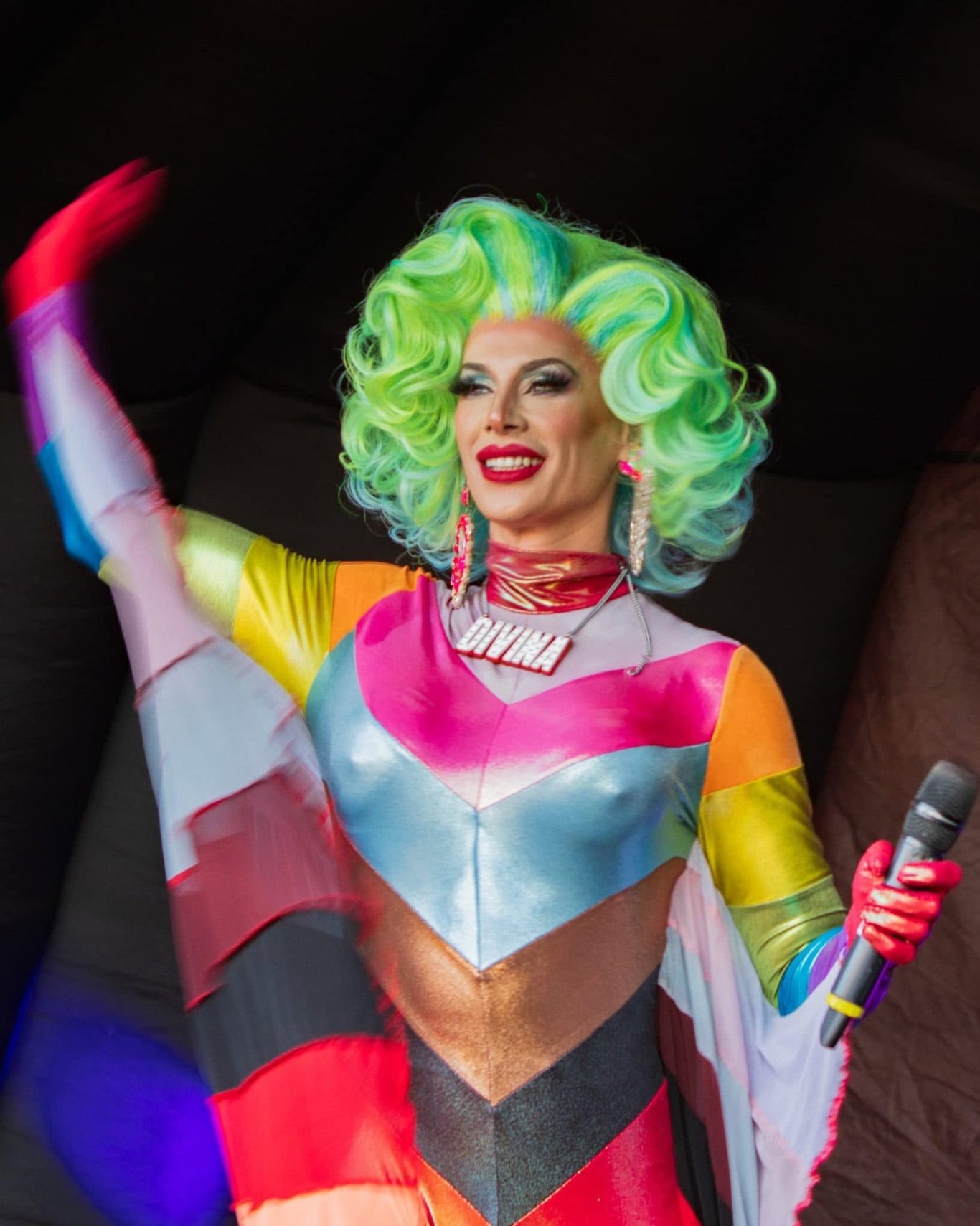 Divina DiCampo - 2024 A performer with vibrant green hair and a colourful outfit gestures while holding a microphone.