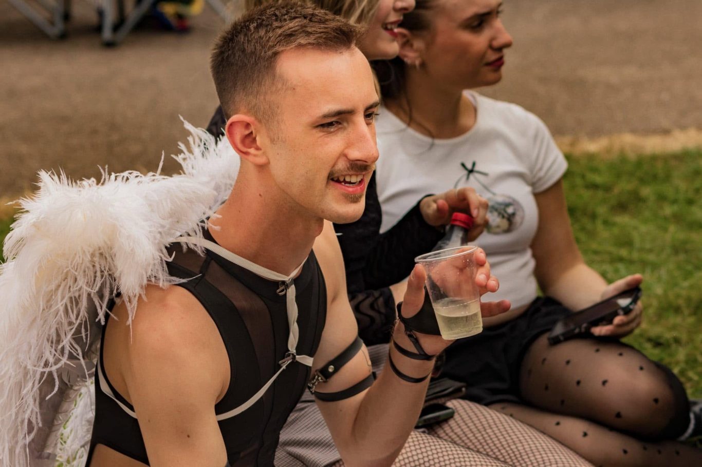 A person in a sheer outfit and wings enjoys a drink, seated beside another individual.