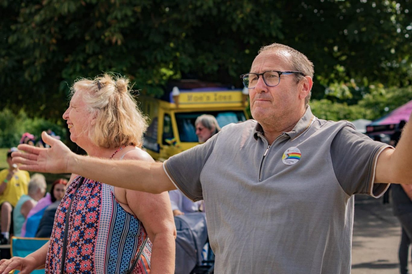 Man with glasses gestures animatedly, while a woman looks on, in a vibrant outdoor setting.