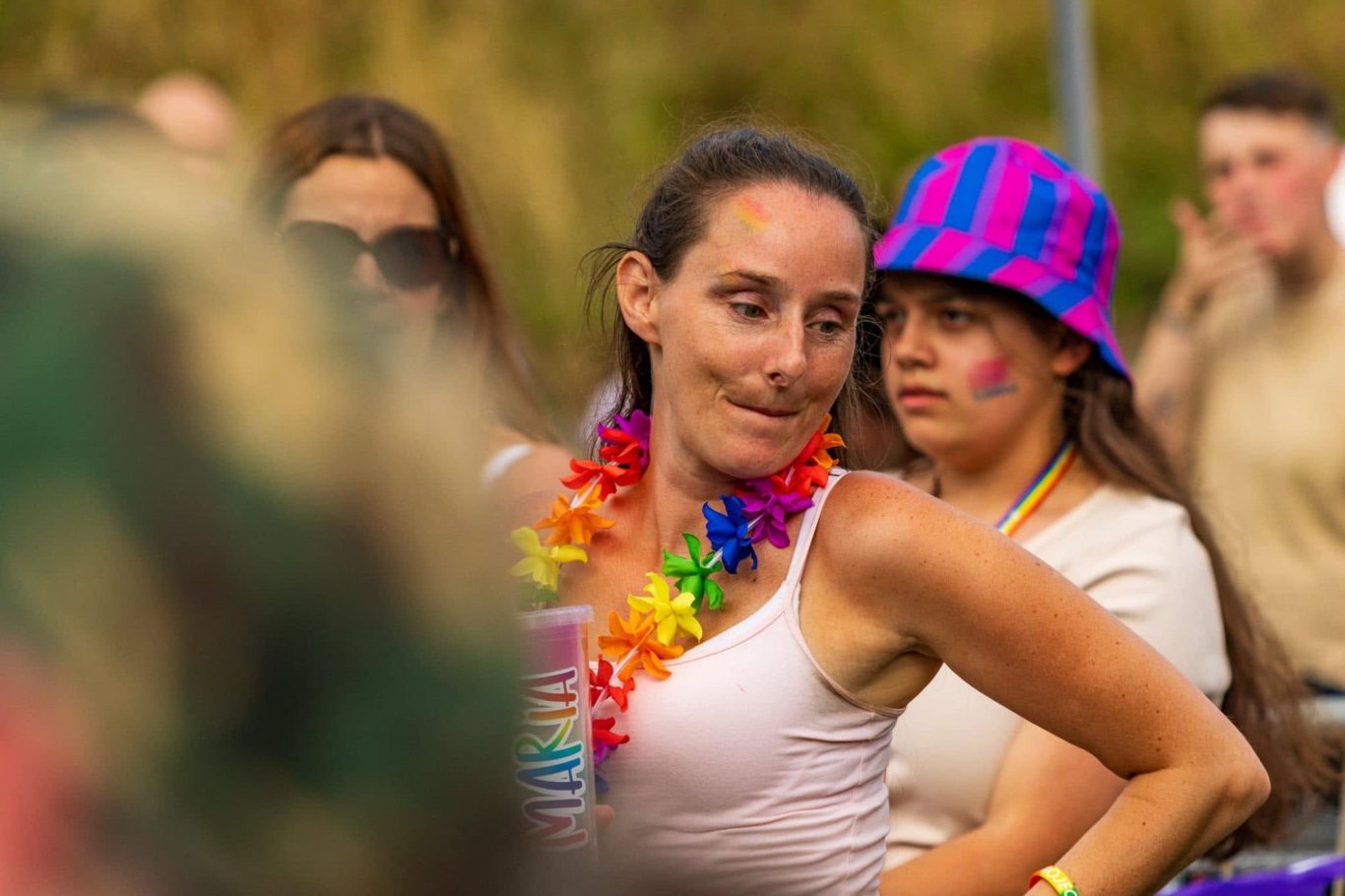 A woman wearing a rainbow lei dances, with others in the background at an outdoor event.