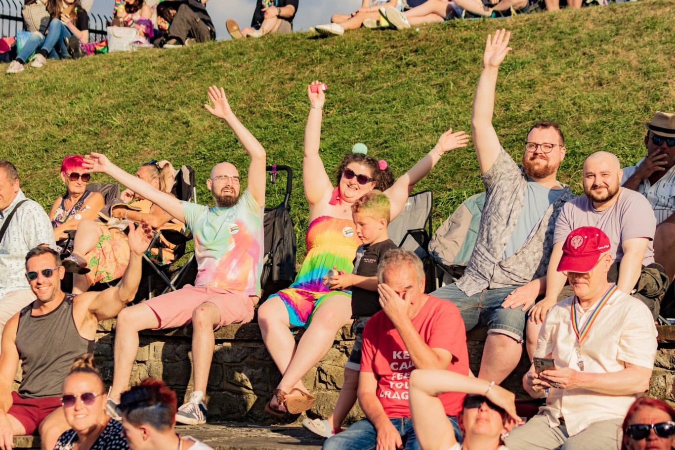 Group of people sitting on grass, joyfully raising their arms and enjoying a sunny day.