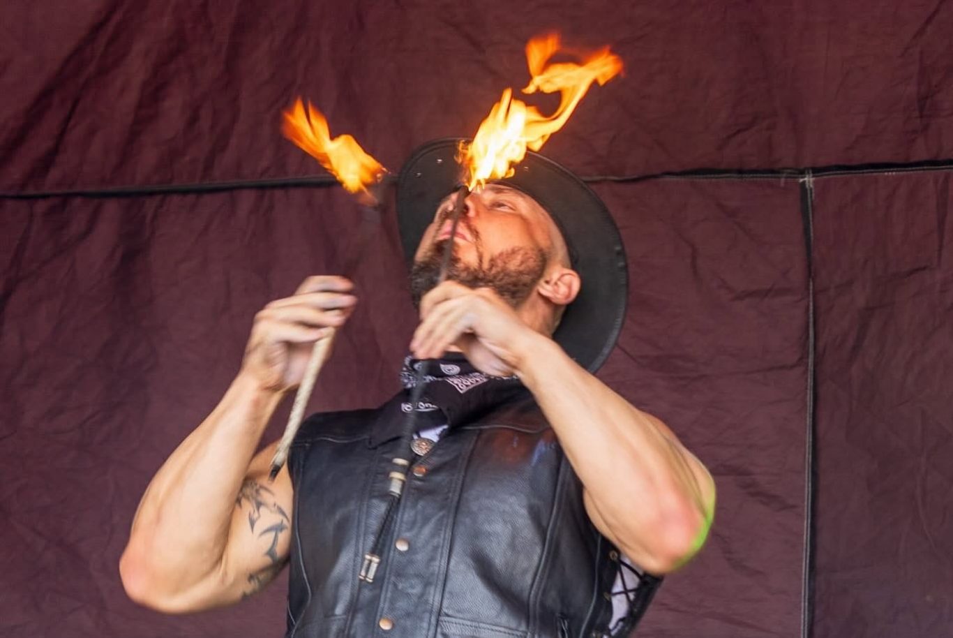 Marshall Arkley - 2024 A performer breathing fire, wearing a black vest and hat, against a dark background.