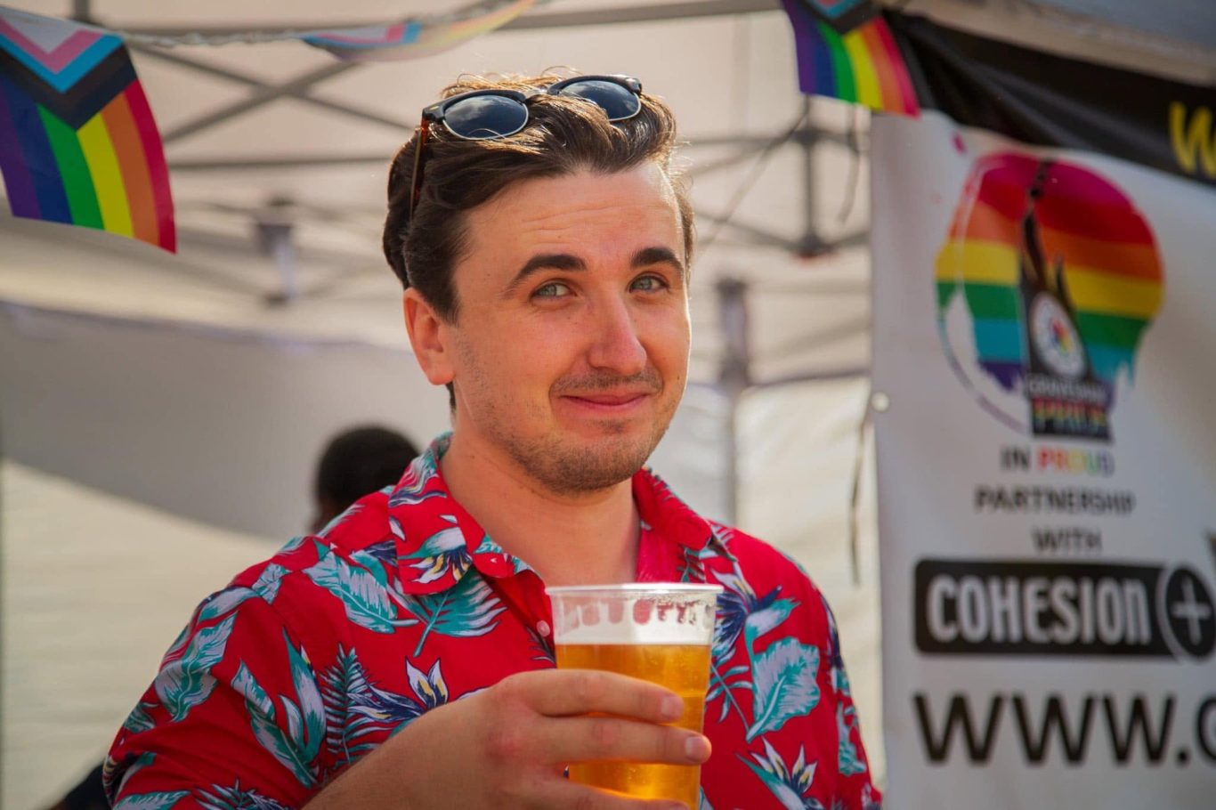 Young man in a floral shirt holding a pint of beer at a pride event with rainbow flags.