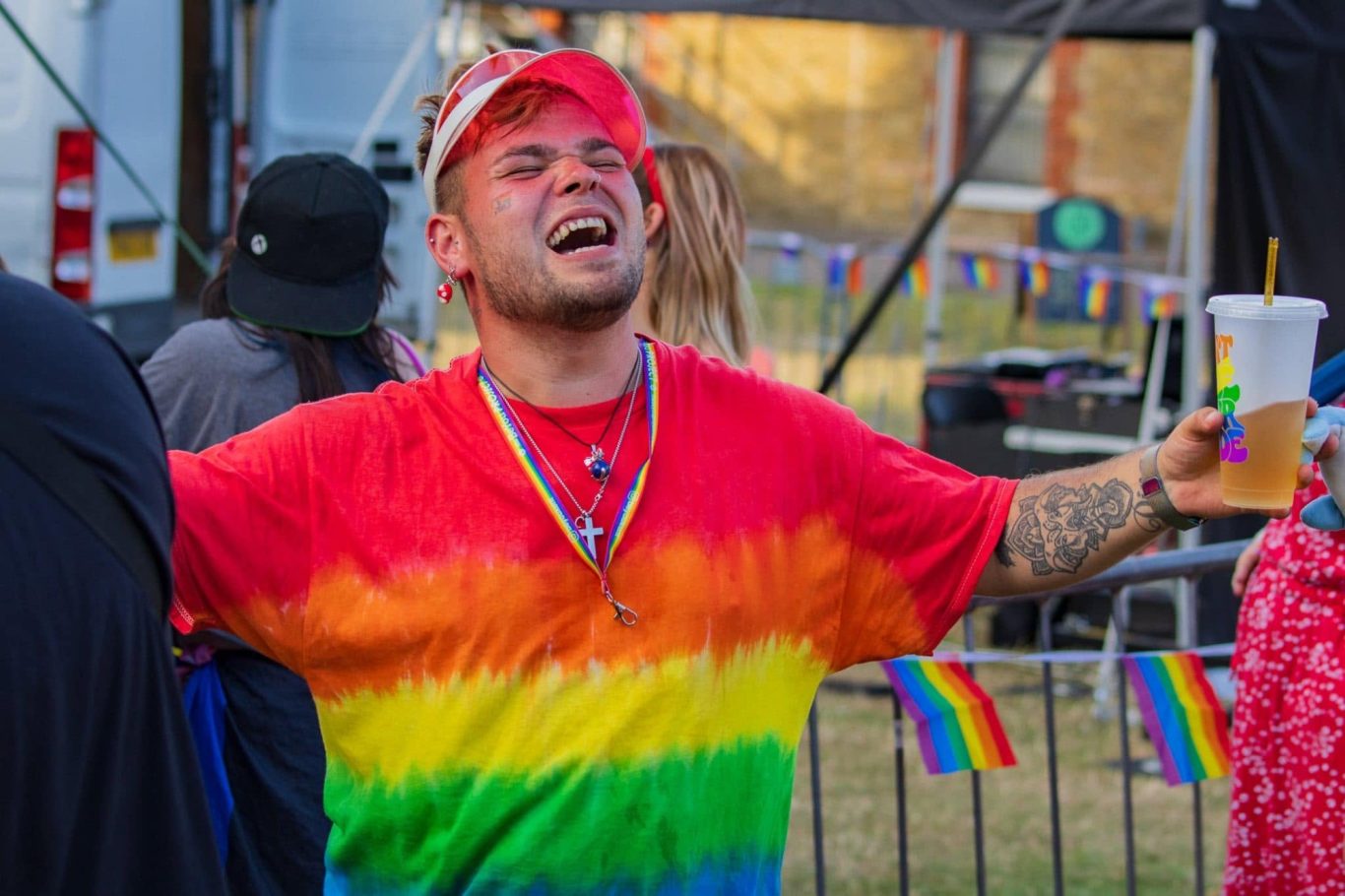 Person in a rainbow t-shirt celebrating, holding a drink at a festive outdoor event.