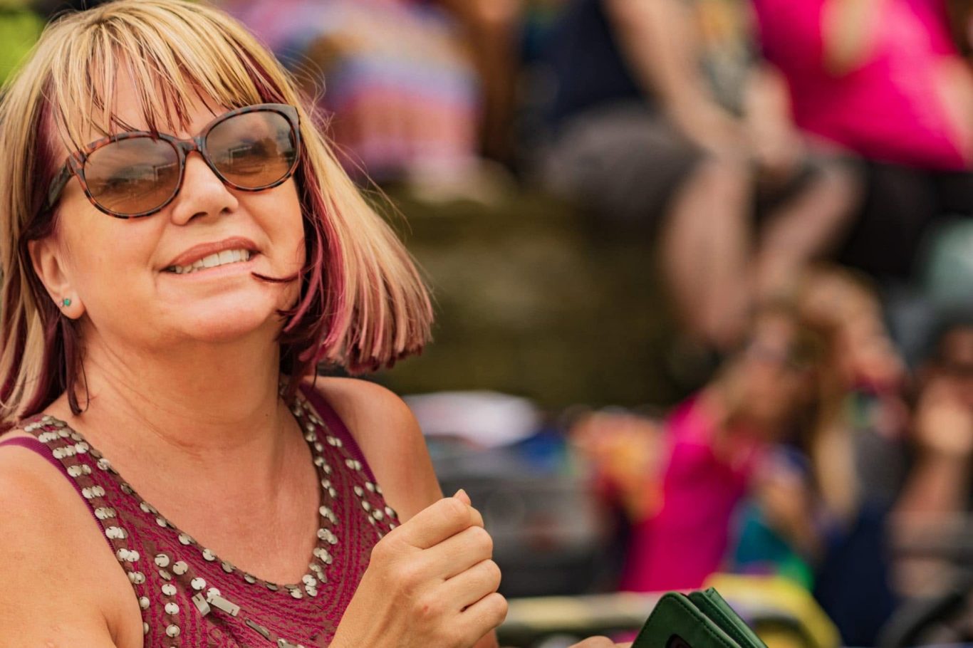 Smiling woman with pink-streaked hair wearing sunglasses, enjoying an outdoor event.