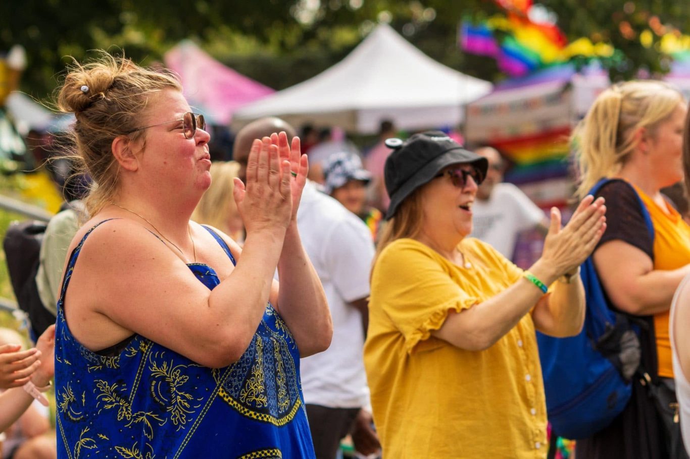 Two women, one in a blue dress and one in a yellow top, applaud at a lively outdoor event.