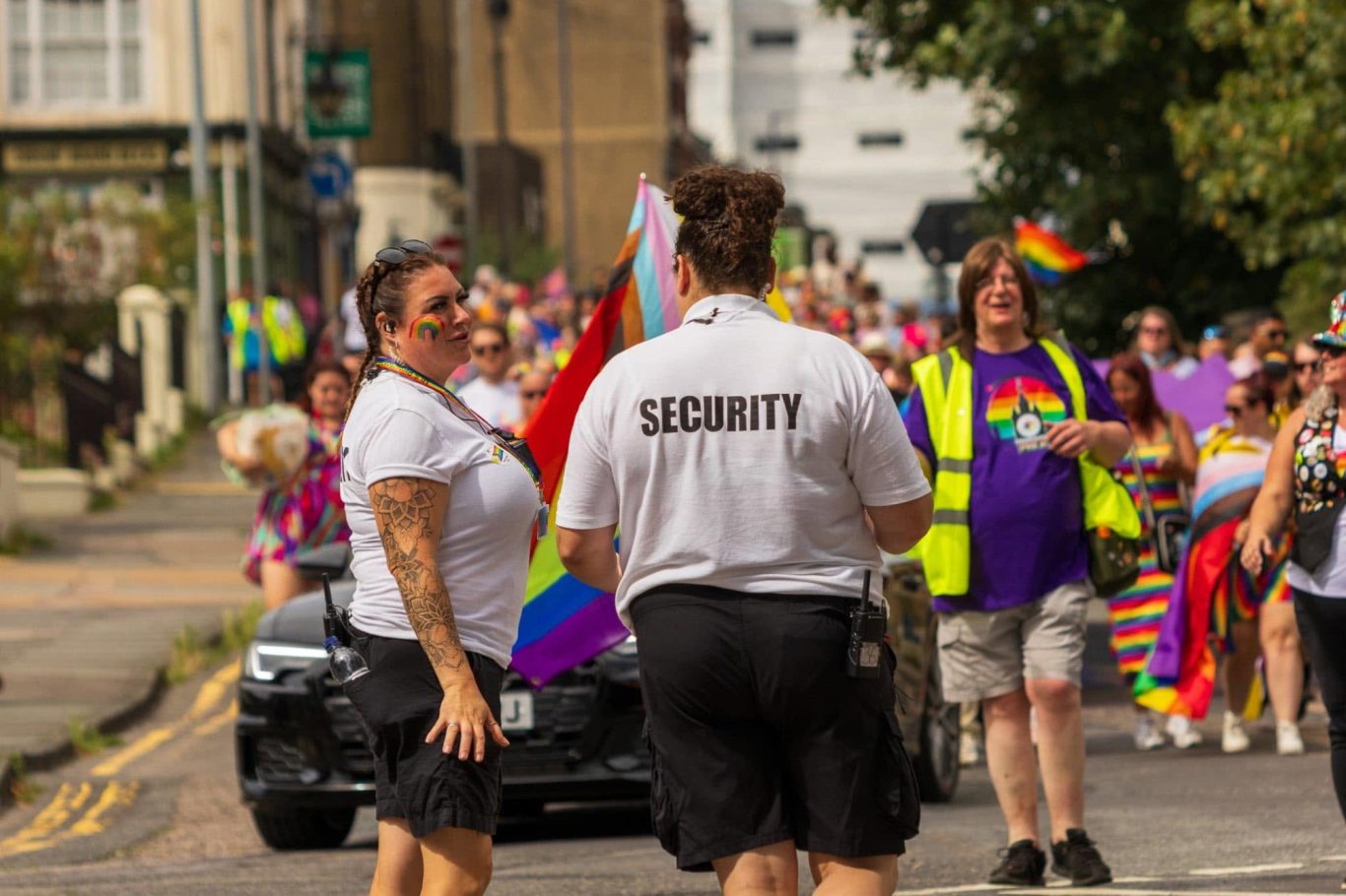 Crowd celebrating Pride parade with security personnel and colourful flags.