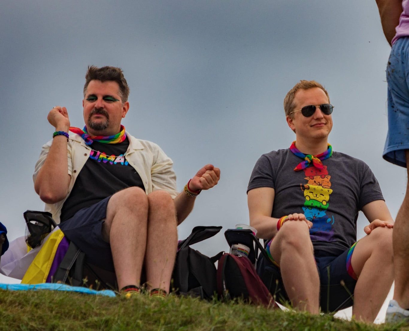 Two men in rainbow accessories sit on a grassy hill, enjoying a festive atmosphere.