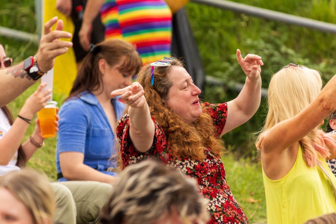 A woman with curly hair gesturing joyfully at an outdoor event, surrounded by others.