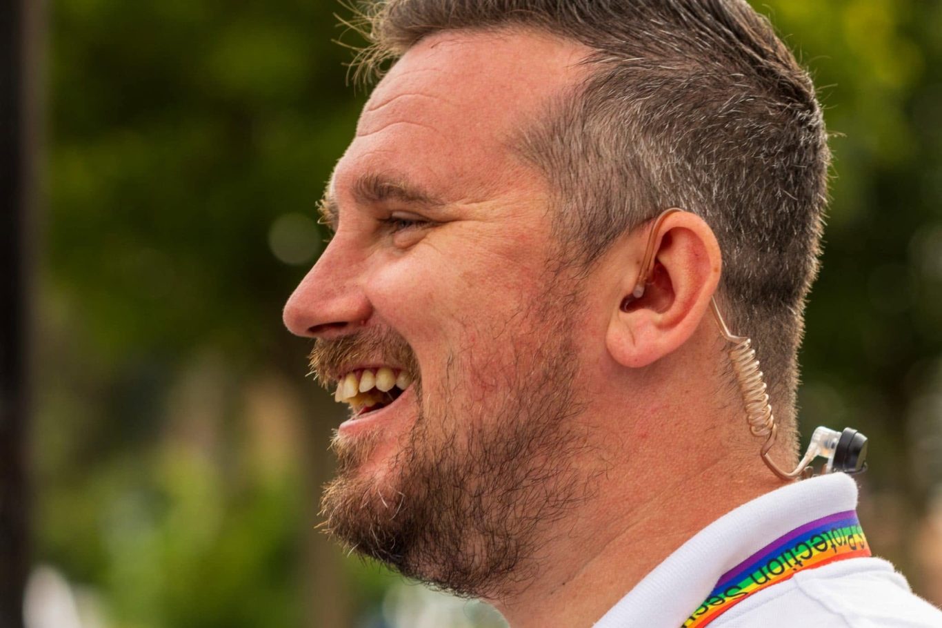 Smiling man with short hair, wearing a rainbow lanyard, outdoors with greenery in the background.