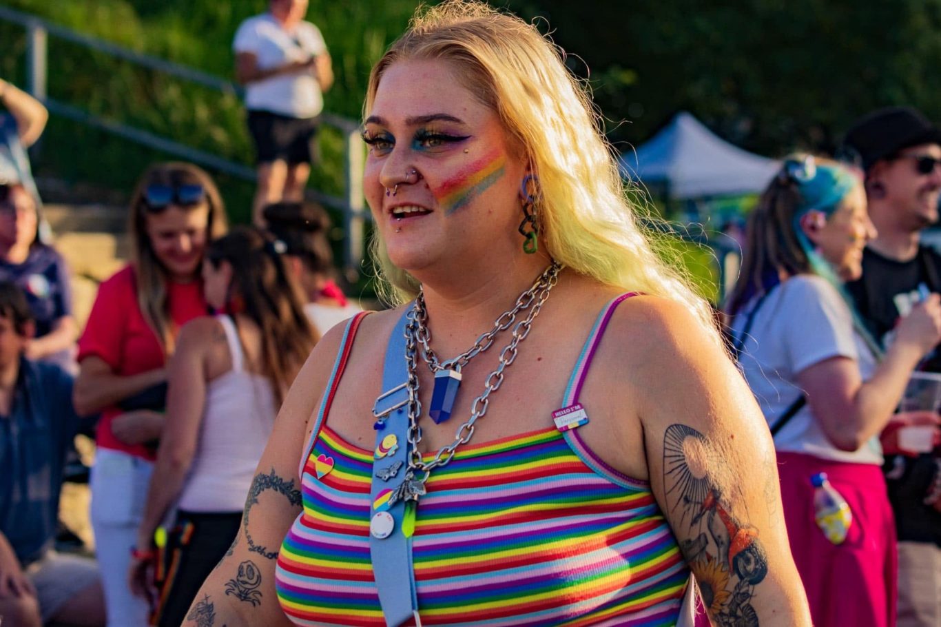 A person with rainbow face paint wearing a striped tank top, surrounded by a lively crowd.